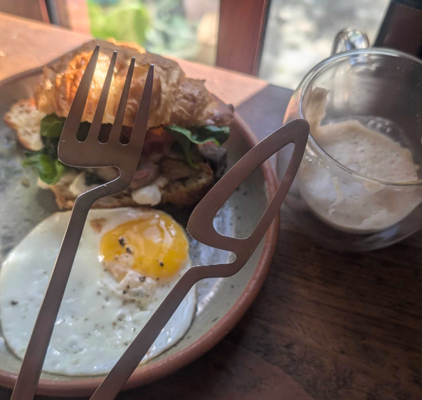 A decoratively attractive set of cutlery with a hole in the middle of the knife, hovering over an egg and pesto croissant next to a mostly finished espresso coffee